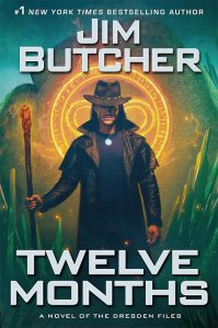 Book cover artwork. A dark-haired man in a fedora and long leather coat stands facing forward, holding a wooden staff topped with a glowing ember. A bright, circular magical sigil with runes glows behind his head like a halo, casting gold light. Green crystal formations rise around him in a shadowy, cavern-like setting. Bold white text reads “Jim Butcher” at the top and “Twelve Months” at the bottom, with the subtitle “A Novel of the Dresden Files.”